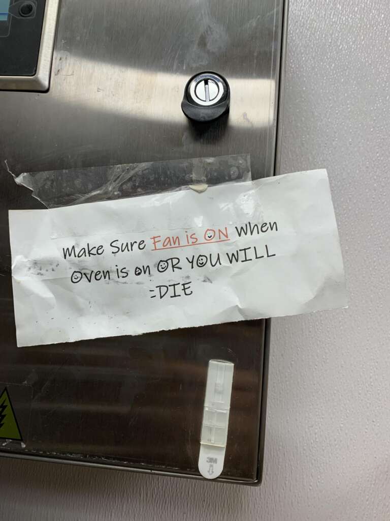 A handwritten sign is taped to a metal surface, likely an oven. The sign reads, "Make sure fan is ON when oven is on or you will die." The text "fan is ON" is highlighted in red. The sign appears slightly crumpled.