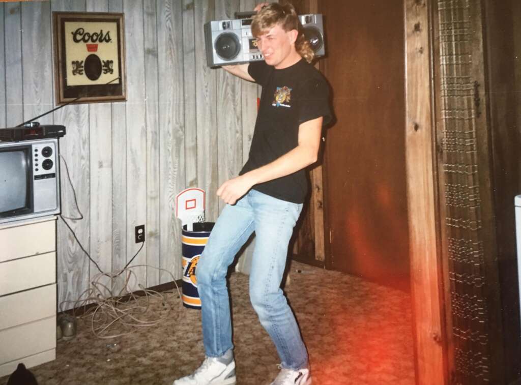 A person in a black t-shirt and blue jeans is holding a boombox on their shoulder while standing in a room with wood-paneled walls. The room has a vintage TV, a Coors sign on the wall, and a mini basketball hoop on a trashcan. The person is smiling and appears to be dancing.