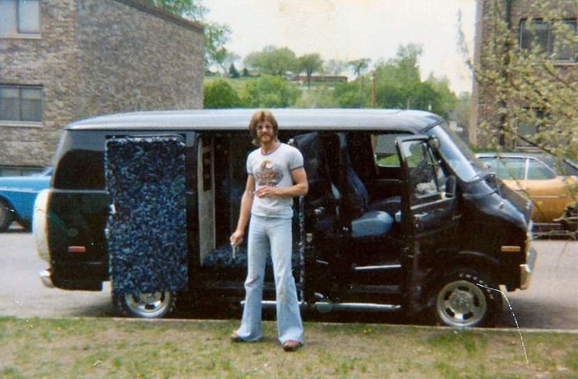 A man with a beard and long hair stands in front of a black van with the side door open. He is wearing a white T-shirt, blue jeans, and sandals. The scene is set in a suburban area with trees, grass, and brick buildings in the background.