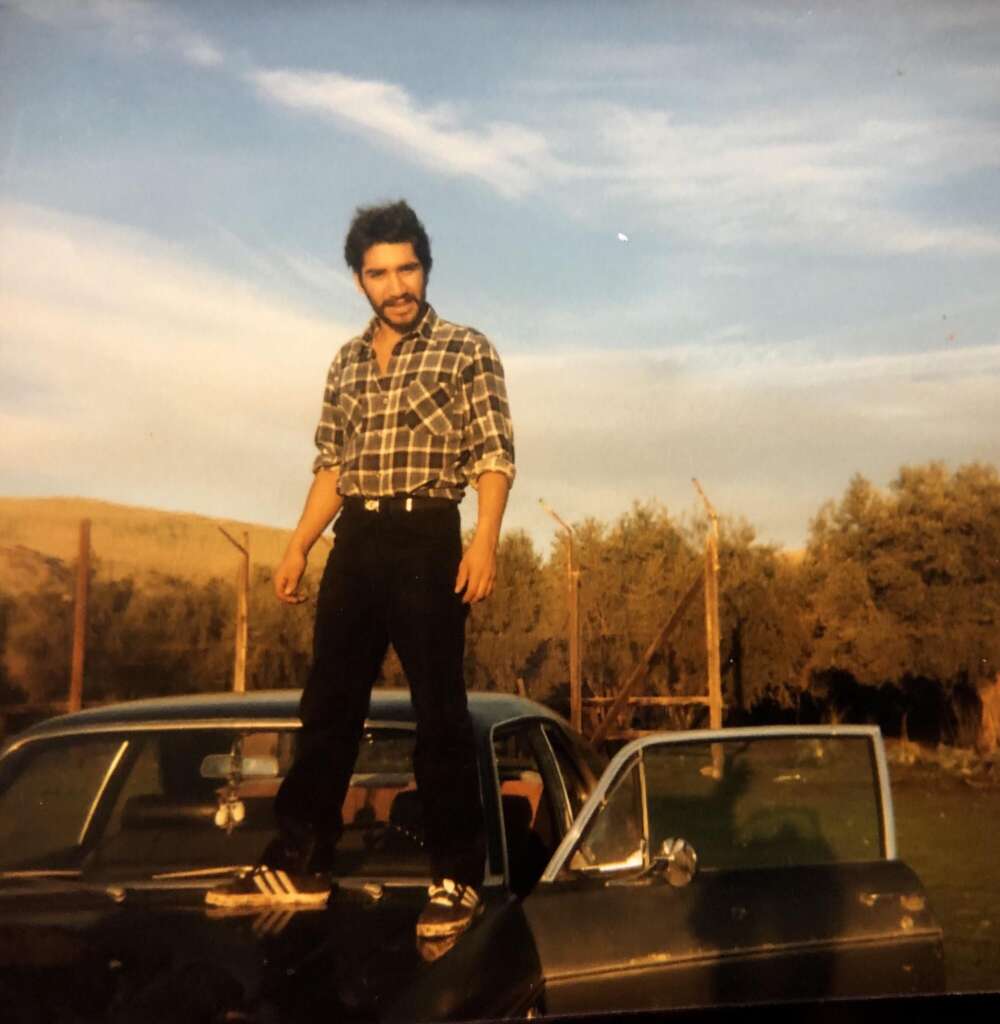 A man with a beard and mustache stands confidently on the hood of a black car with an open driver's side door. He is wearing a plaid shirt, black pants, and sneakers. The background features a fence, trees, and a blue sky with wispy clouds.