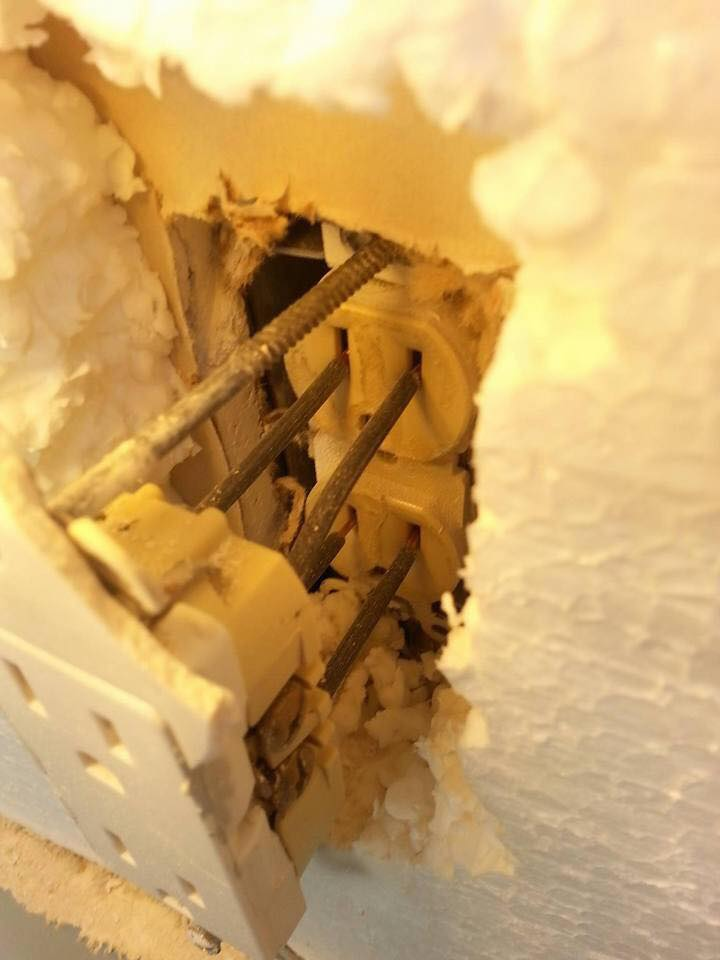 A close-up view of a damaged wall with exposed rebar and electrical components. The wall appears to have been partially cut or broken open, revealing the internal structure and wires. There is a white, foam-like material surrounding the exposed area.