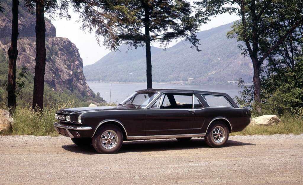 A black classic station wagon is parked on a dirt road surrounded by trees. In the background, there is a large body of water and hills or mountains. The car's design suggests it is from the mid-20th century.