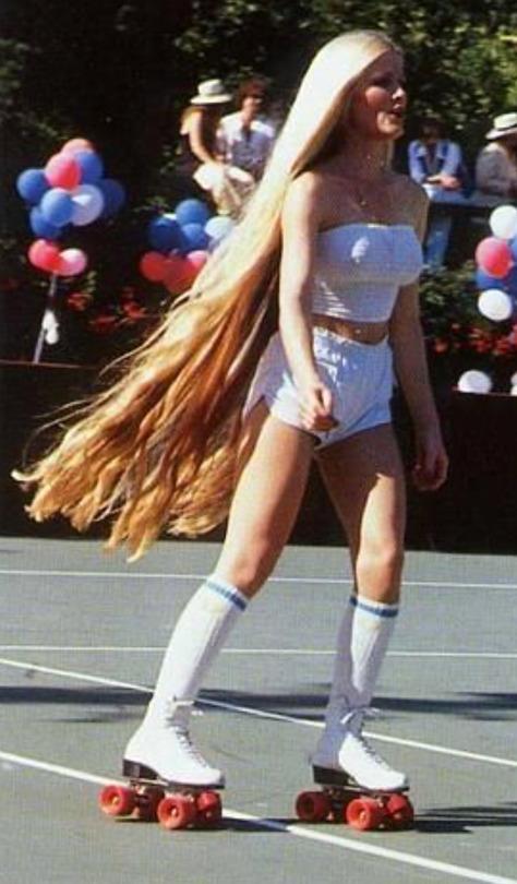 A person with long, flowing blonde hair roller skates on a tennis court wearing a white tube top, high-waisted shorts, and knee-high socks. The background features red, white, and blue balloons and spectators sitting on benches.