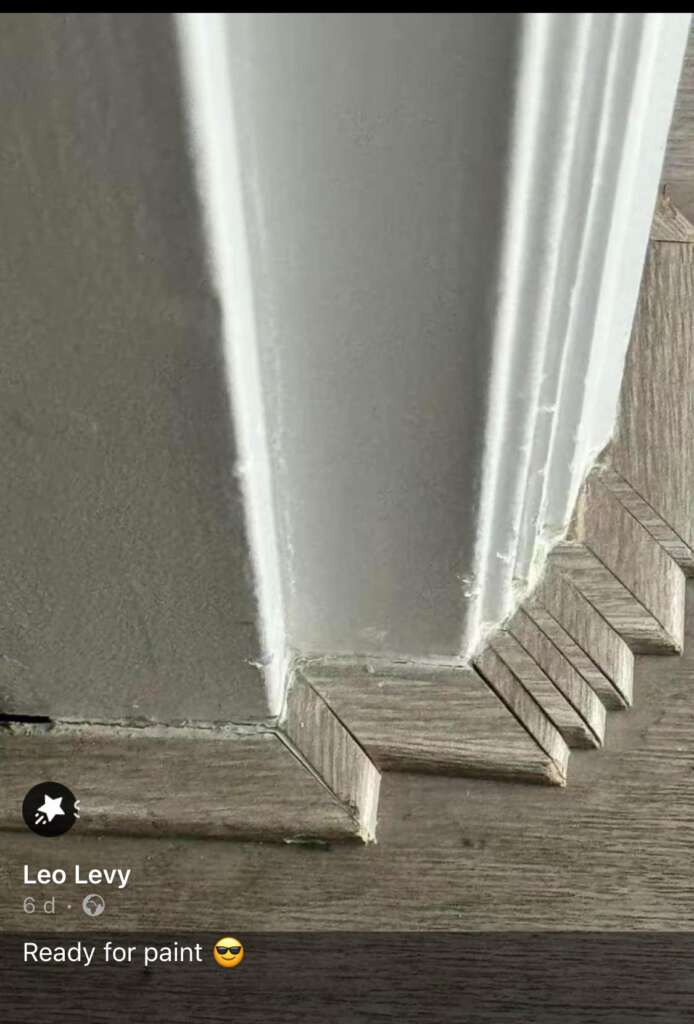 A close-up of the base of a wall's corner, showing detailed and precise flooring work with small, angled pieces of wood neatly fitted together. The wall trim above the flooring appears prepped for painting. A social media post by Leo Levy, text says "Ready for paint 😎".