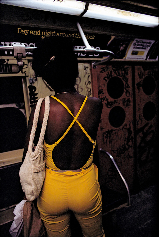 A person stands in a subway car, facing away from the camera. The person is wearing a bright yellow outfit with crisscross straps, and carrying a beige tote bag over their shoulder. The subway walls are covered with graffiti, and an overhead sign reads "Day and night around the clock.