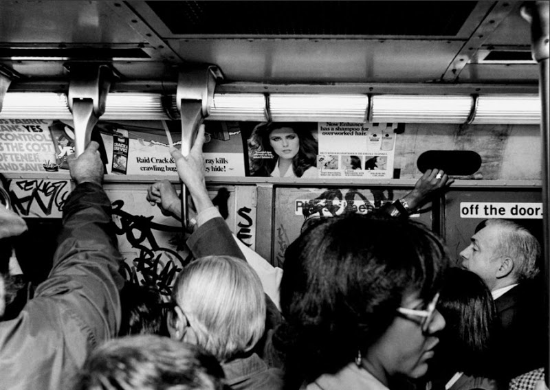 A crowded subway car with numerous passengers standing, holding onto overhead handrails. The walls are covered with graffiti and posters. One ad features a woman's face with the headline, "Real Crack Heros." People are tightly packed, engrossed in the commuter experience.