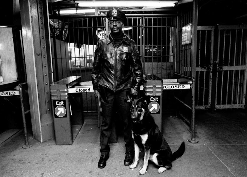 A uniformed police officer stands next to a German Shepherd police dog at a subway entrance. The officer wears a leather jacket and cap, and the dog sits alertly by his side. Signs indicating "Exit" and "Closed" can be seen behind them.