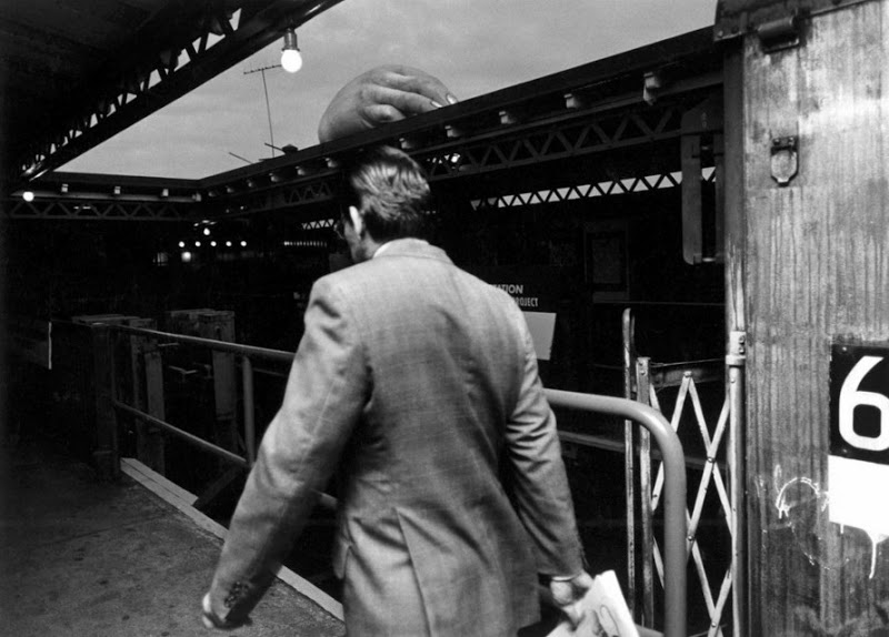 A person in a suit is seen from behind walking into a tunnel-like structure. The top of a large inflatable structure is visible over the roof of the tunnel. The person is holding a rolled-up newspaper with the number 66 visible on the right side of the image.