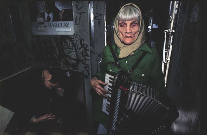 An elderly woman is playing an accordion while wearing a green coat and a beige headscarf inside a graffiti-covered room. Another person, wearing dark clothing, is sitting and leaning against the wall in the background. A "BARCLAY" poster is partially visible.