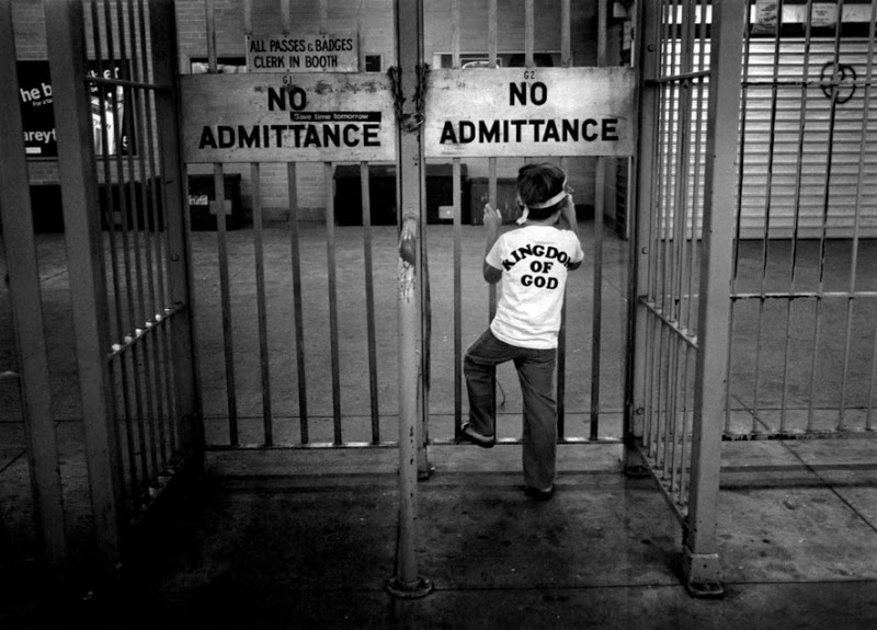 Black and white image of a child wearing a "Kingdom of God" t-shirt, climbing metal bars in front of two gates labeled "NO ADMITTANCE." A sign above the gates reads "ALL PASSES, BADGES CLEAR IN BOOTH." The setting appears to be industrial or institutional.