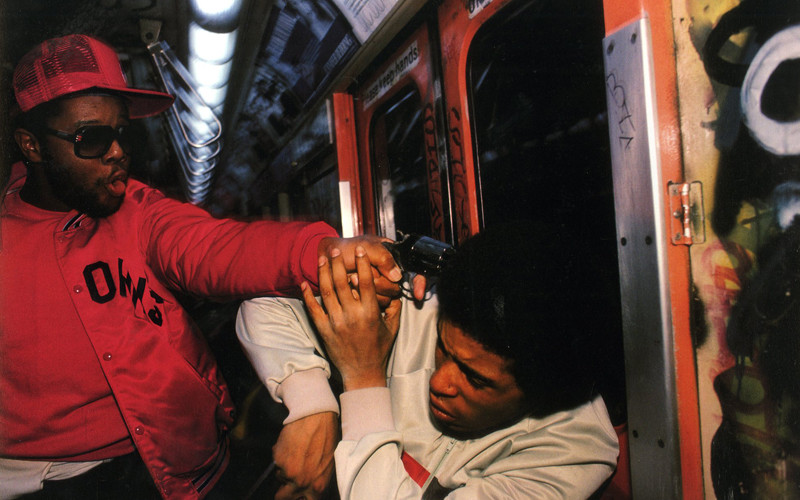 In a dimly-lit subway car, an individual in a red jacket and cap points a gun at another person's head, who appears terrified and is shielding themselves with their hands. The subway walls are covered in graffiti.