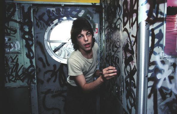 A person with medium-length hair wearing a light-colored t-shirt is standing in a graffiti-covered space, possibly a subway car. They are holding onto a surface with their left hand and looking towards the camera with a surprised or startled expression.