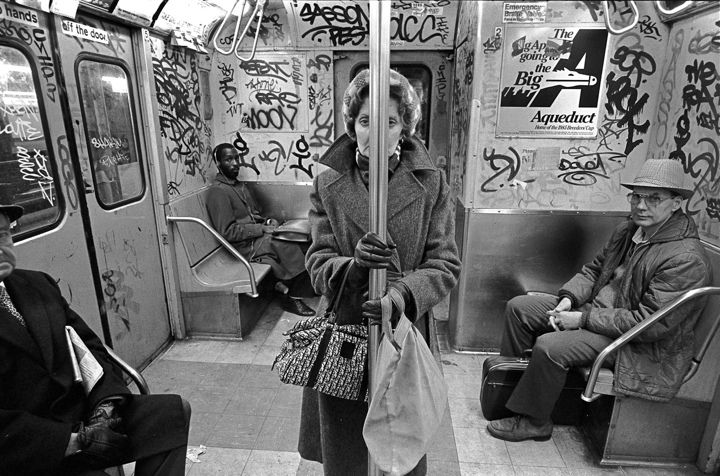 Black and white photo of a subway car interior with graffiti-covered walls. Four passengers are visible; a woman standing in the center holding a pole, two men seated on the right, and another seated on the left. An Aqueduct advertising poster is on the wall.