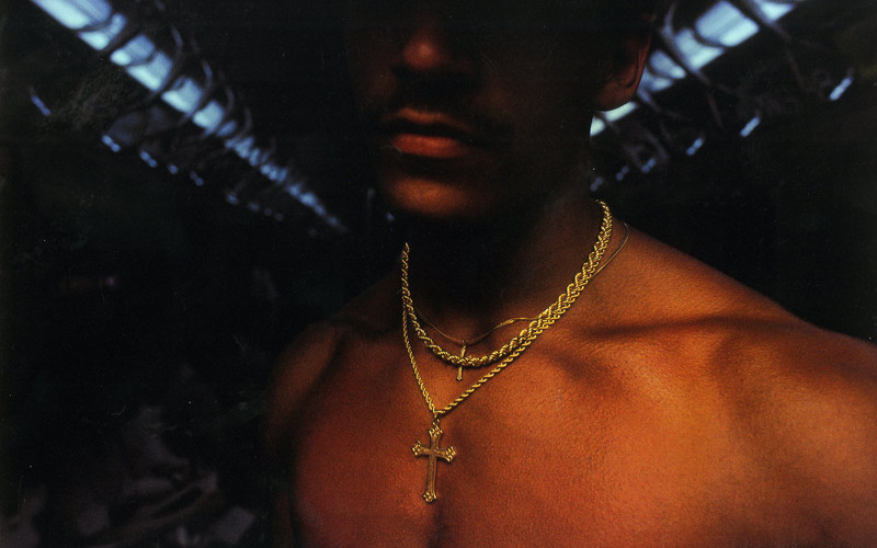 A shirtless person is shown from the shoulders up, wearing multiple gold chains around their neck, including one with a cross pendant. The background is dimly lit with a row of overhead lights visible.