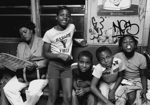 A black-and-white photo of four young boys and a woman on a subway. The boys are smiling and posing for the camera, while the woman sits beside them, reading a magazine. The subway walls are covered with graffiti.