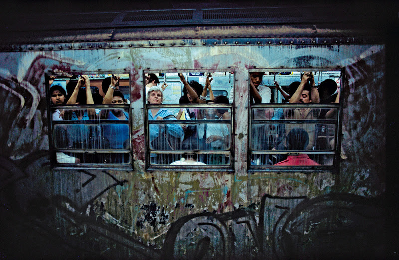 Passengers tightly packed inside a graffiti-covered subway car, viewed through the windows. Their expressions are varied, and many are holding onto the overhead handrails for support. The image captures the crowded and gritty atmosphere of urban public transportation.