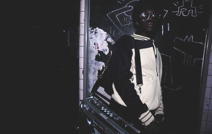 A person wearing a black and white jacket, glasses, and a hat stands against a dark wall with graffiti. They are holding a large boombox. The scene is dimly lit with a street art vibe.