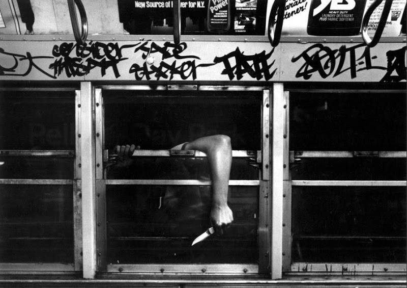 A black-and-white photo shows a person inside a graffiti-covered bus or train. The person's arm is extended through the window, holding a knife. The scene is tense and edgy, suggesting a moment of potential violence or threat.