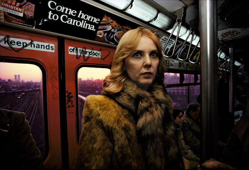 A woman with blonde hair, wearing a fur coat, stands inside a graffiti-covered subway car. The orange doors show a view of the city at sunset, while an advertisement above reads "Come home to Carolina." Other passengers are seated in the background.