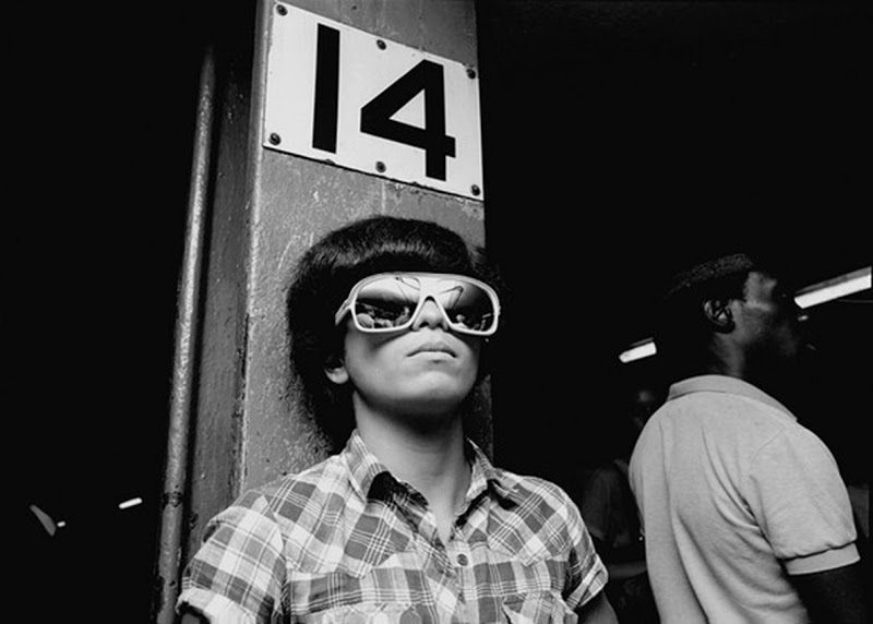 A person wearing large sunglasses leans against a pole labeled "14." The person is dressed in a checkered shirt and appears relaxed. Another individual, partially visible, stands in the background. The scene is in black and white.