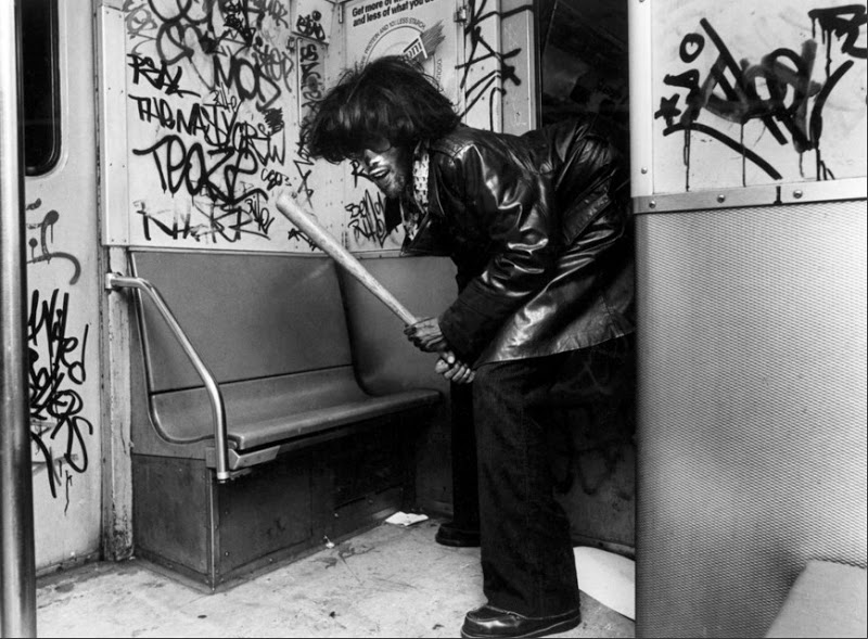 A person holding a baseball bat in a subway car with graffiti-covered walls, wearing a dark leather jacket. The individual is bent over and facing the camera, the subway scene appears gritty and urban.