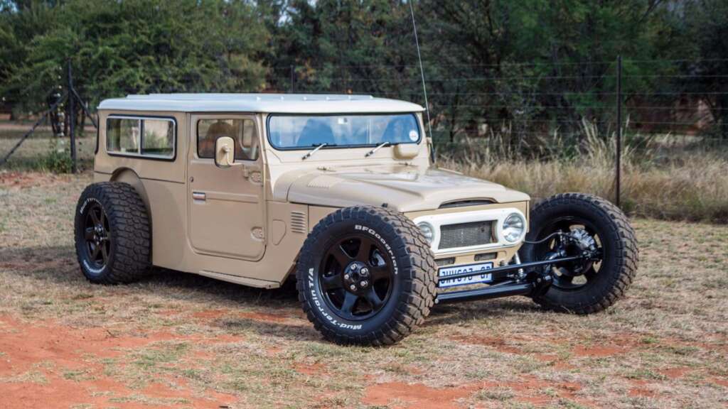 A custom beige hot rod vehicle with a vintage, rugged design is parked on a grassy area. It has large off-road tires, a modified front grille, and an overall retro aesthetic. Trees and a wire fence are visible in the background.