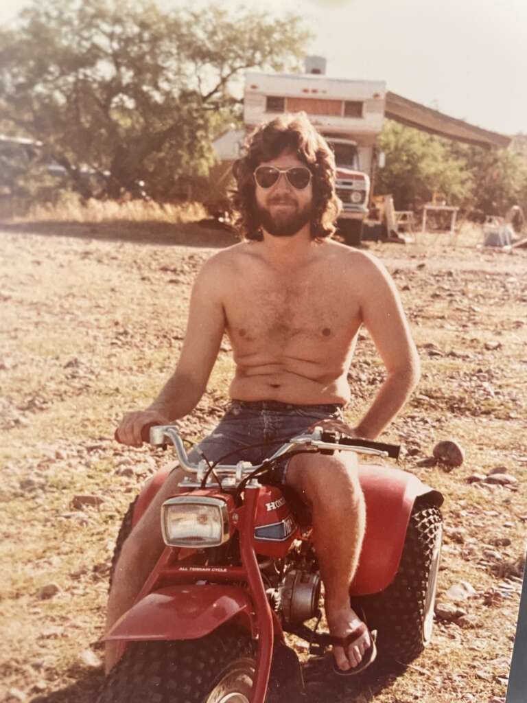 A bearded man wearing sunglasses, denim shorts, and sandals sits on a red three-wheeler in an outdoor area with a camper and trees in the background. The atmosphere appears warm and sunny.