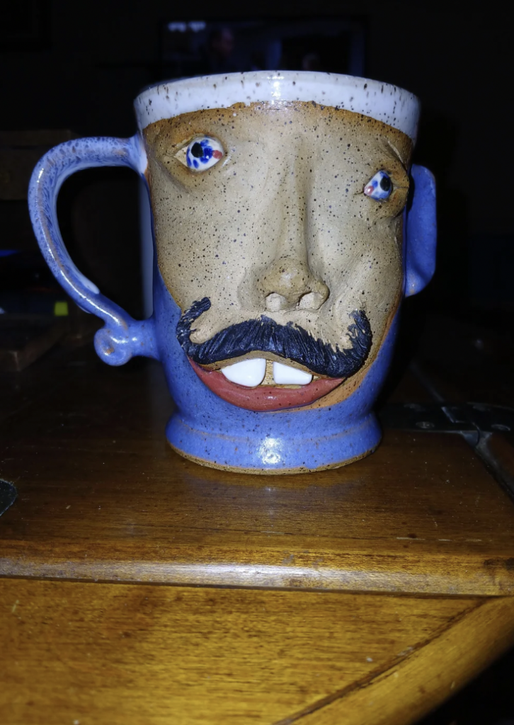 A ceramic mug with a whimsical face design, featuring a protruding nose, mustache, and two eyes that appear to be made of blue and white beads. The handle connects to one ear, and the mug has a blue glaze on the exterior. It sits on a wooden surface.
