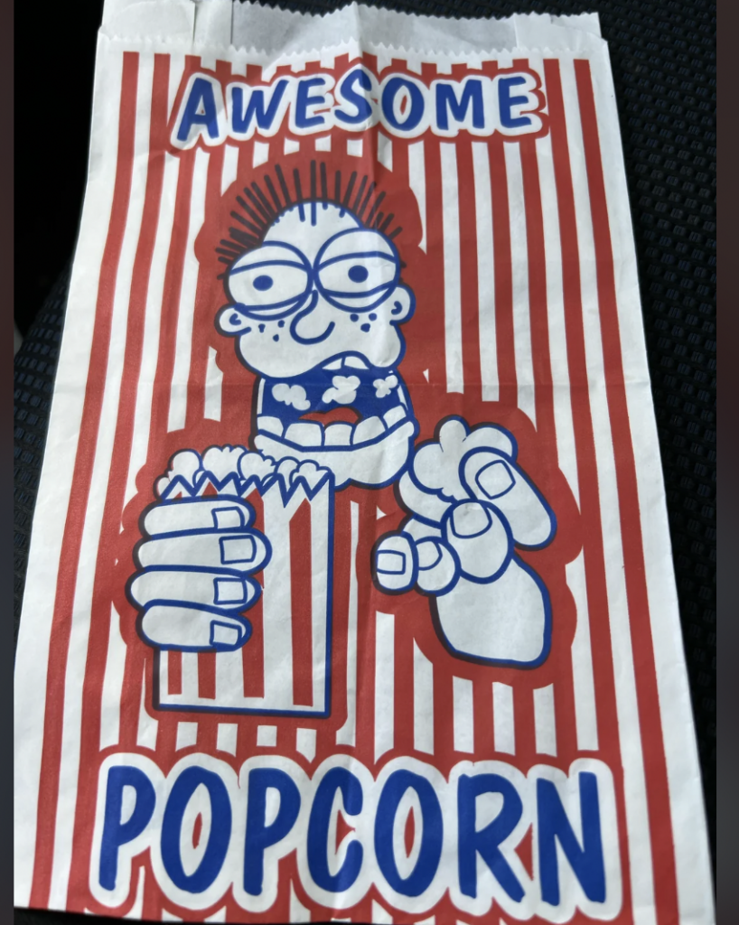 A white and red striped popcorn bag with a cartoon character holding a popcorn bucket and giving a thumbs-up. The cartoon character has spiky hair, glasses, and exaggerated facial features. The words "AWESOME POPCORN" are written at the top and bottom of the bag.