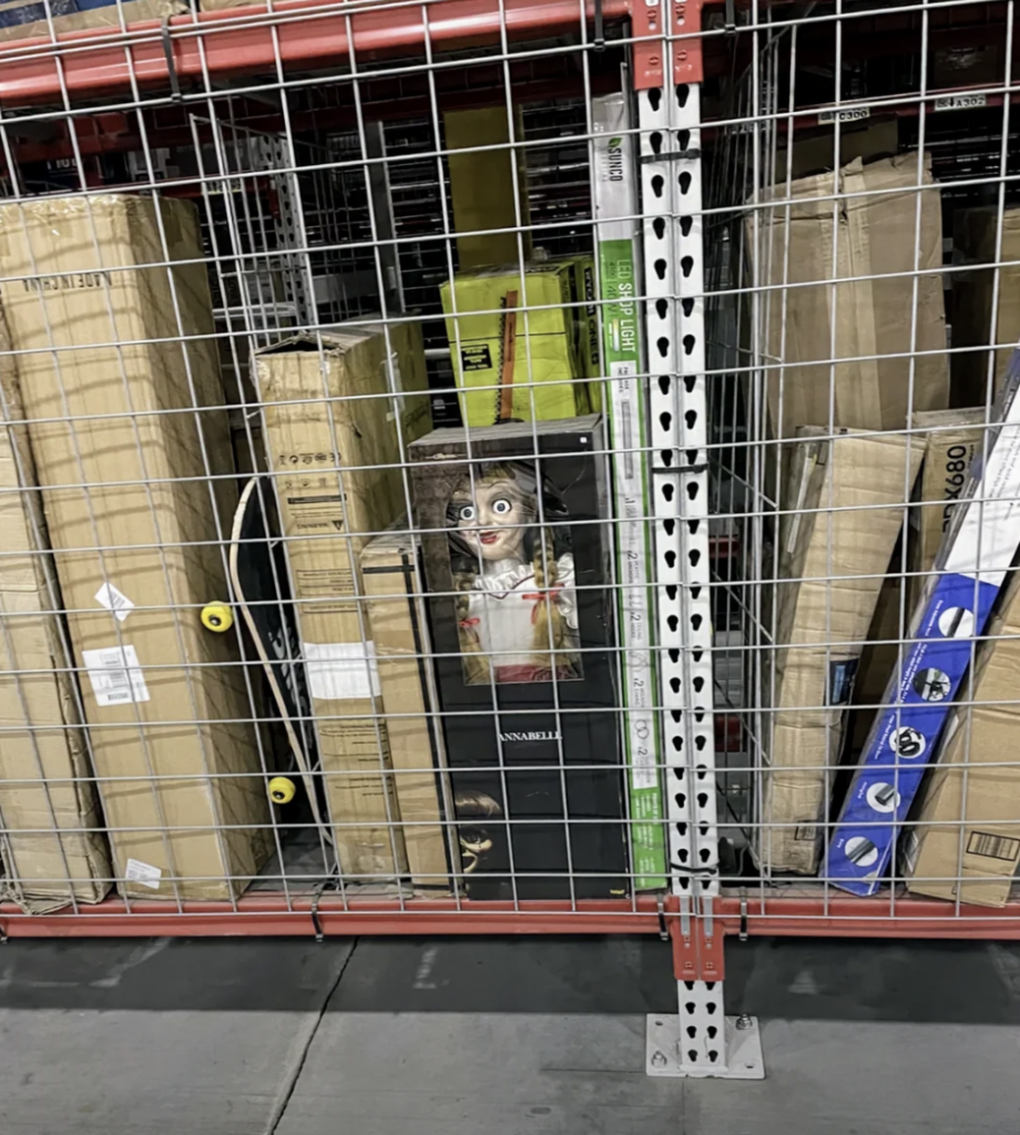 A large metal storage shelf in a warehouse contains various large boxes and a life-sized creepy doll with wide, staring eyes and a distressed expression, partially hidden among them, visible through the shelf's metal grid. The floor is concrete.