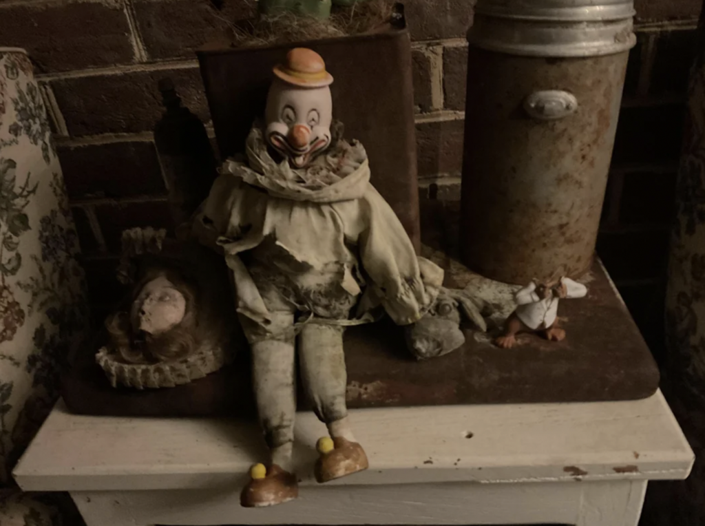 A worn-out clown doll, with a tattered outfit and a hat, sits on a shelf against a brick wall. Beside it lies an old doll's head with a partially cracked face and an antique cylindrical container. The scene is dimly lit, giving it a vintage and eerie ambiance.