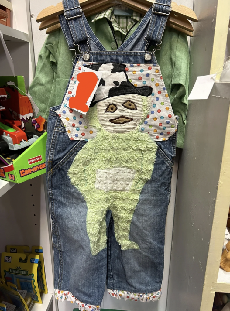 A child's outfit hangs on a rack, featuring denim overalls with a patch of a green character wearing a black and white hat on the front. Underneath the overalls, a green long-sleeve shirt is visible. The outfit is displayed in a store with toys on the nearby shelves.