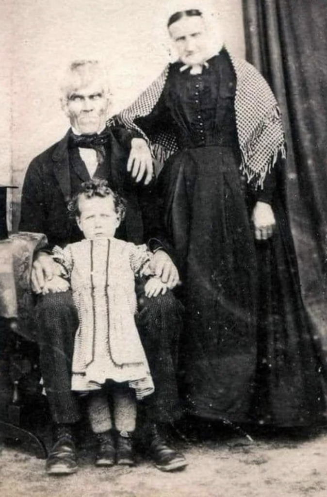 A black and white vintage photograph of three people. An elderly man with white hair and glasses sits holding a young child in a white dress, while an elderly woman in a dark dress and shawl stands beside them with her hand on the man's shoulder.