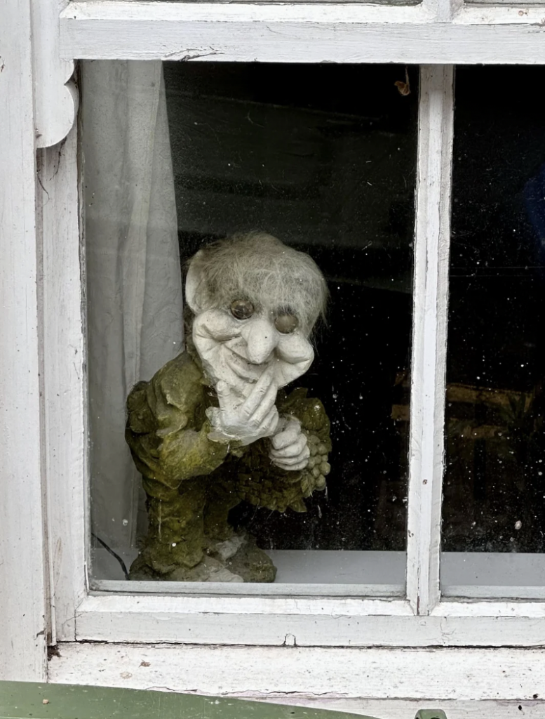 A small, whimsical statue of a creature with a mischievous grin, bushy eyebrows, and tousled gray hair peeks out from behind a window. The creature has exaggerated facial features and is wearing a green outfit. Dust and grime are visible on the window.