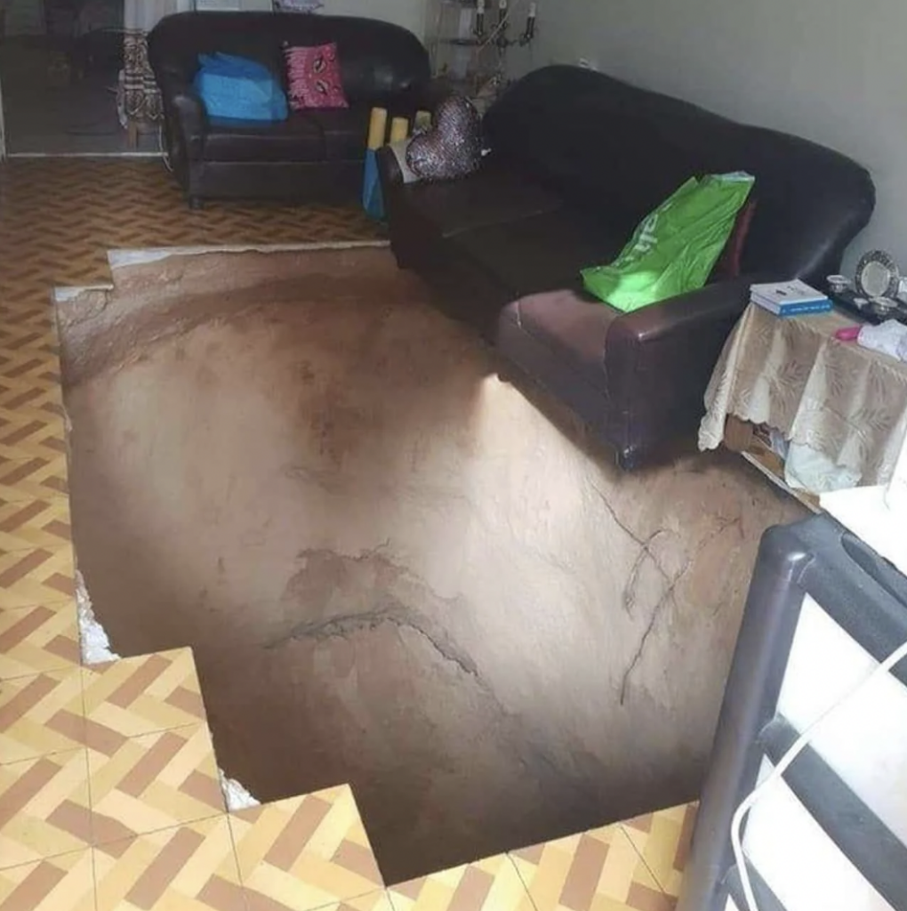 A living room with a large sinkhole spanning most of the floor, partially exposing the ground beneath. The sinkhole is directly in front of two sofas, which remain intact. The room also has tiled flooring with a geometric pattern surrounding the sinkhole's edge.