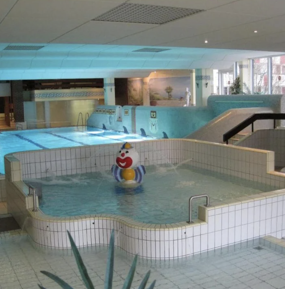 An indoor swimming pool complex with a main pool in the background and a small, separate shallow pool in the foreground featuring a clown-themed inflatable toy. The area is well-lit with overhead lights and has white tiled walls and floors.