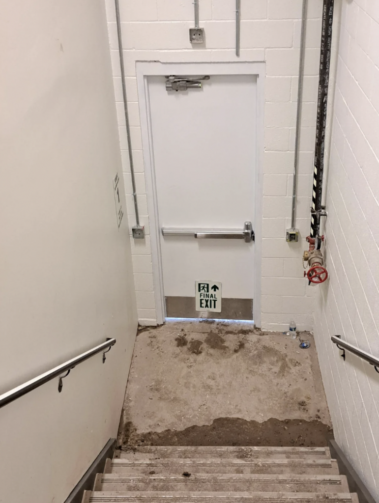 A stairwell leads down to a white door marked "Final Exit" with a green sign. The stairs and floor are dirty and partially covered with debris. The walls are painted white, and various pipes and electrical conduits run along them.
