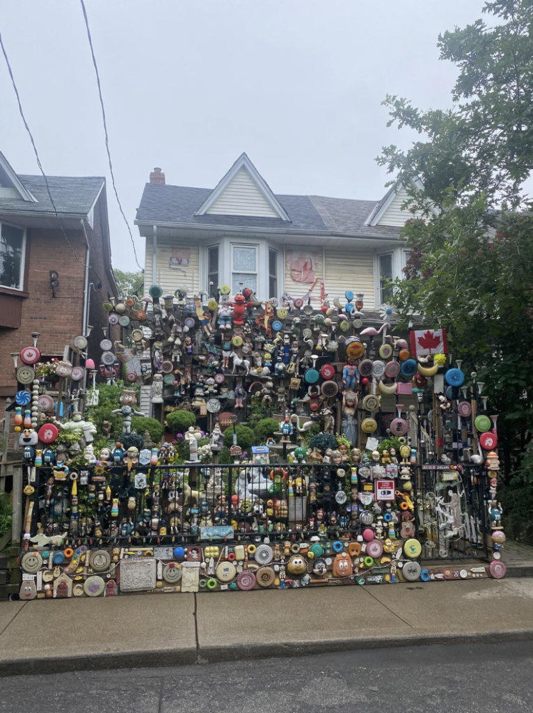A house with its front yard and fence covered in various colorful decorations, including signs, toys, and ornaments, creating a densely packed, vibrant display. The fence exhibits an eclectic mix of objects, and a Canadian flag is visible among the decorations.