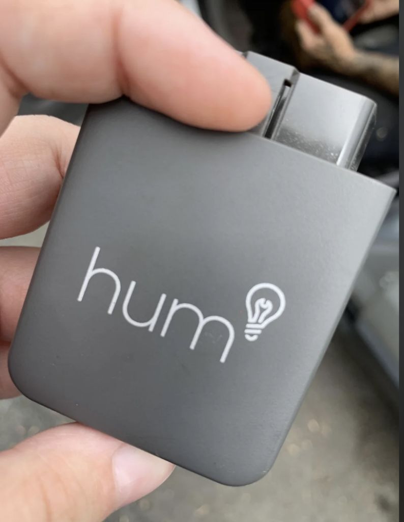A hand holding a small rectangular gray device with the word "hum" and a light bulb icon printed on it. The background is slightly blurred, showing parts of a car interior.