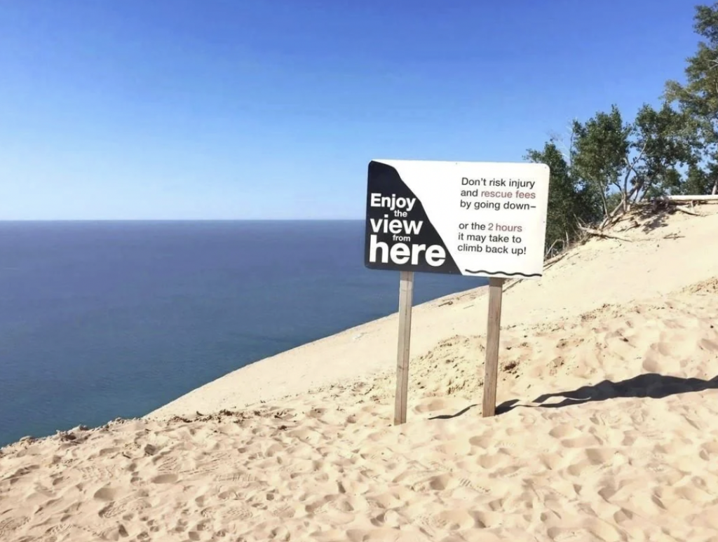 A warning sign on a sandy hill overlooking a vast body of water. The sign reads, "Enjoy view from here. Don't risk injury and rescue fees by going down–or the 2 hours it may take to climb back up!" Nearby are a few trees and shrubs. The sky is clear and blue.