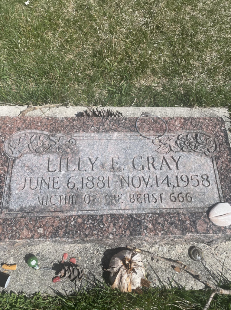 Image of a gravestone for Lilly E. Gray, dated June 6, 1881 – November 14, 1958. The inscription below her name reads "Victim of the Beast 666." There are small items and decorations placed at the base of the gravestone.