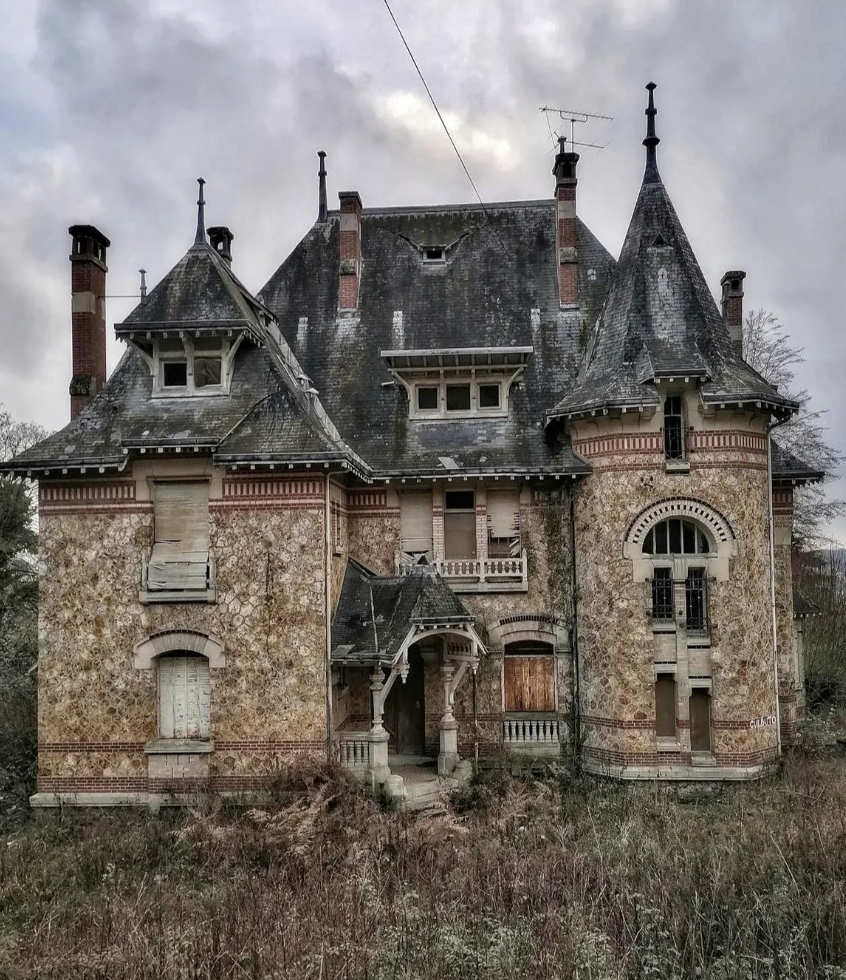 An old, abandoned mansion with boarded-up windows and overgrown vegetation. The building has a steep, slate roof with multiple chimneys and tall, pointed turrets. The cloudy, overcast sky adds to the eerie, neglected atmosphere.