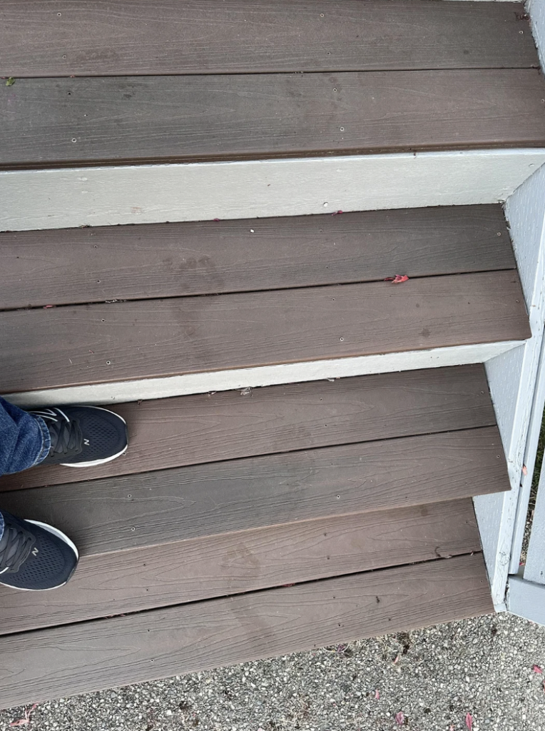A person wearing black sneakers and blue jeans stands on the edge of brown wooden stairs next to a white railing. The stairs have some scattered small debris, and the surrounding area appears to be gravel.