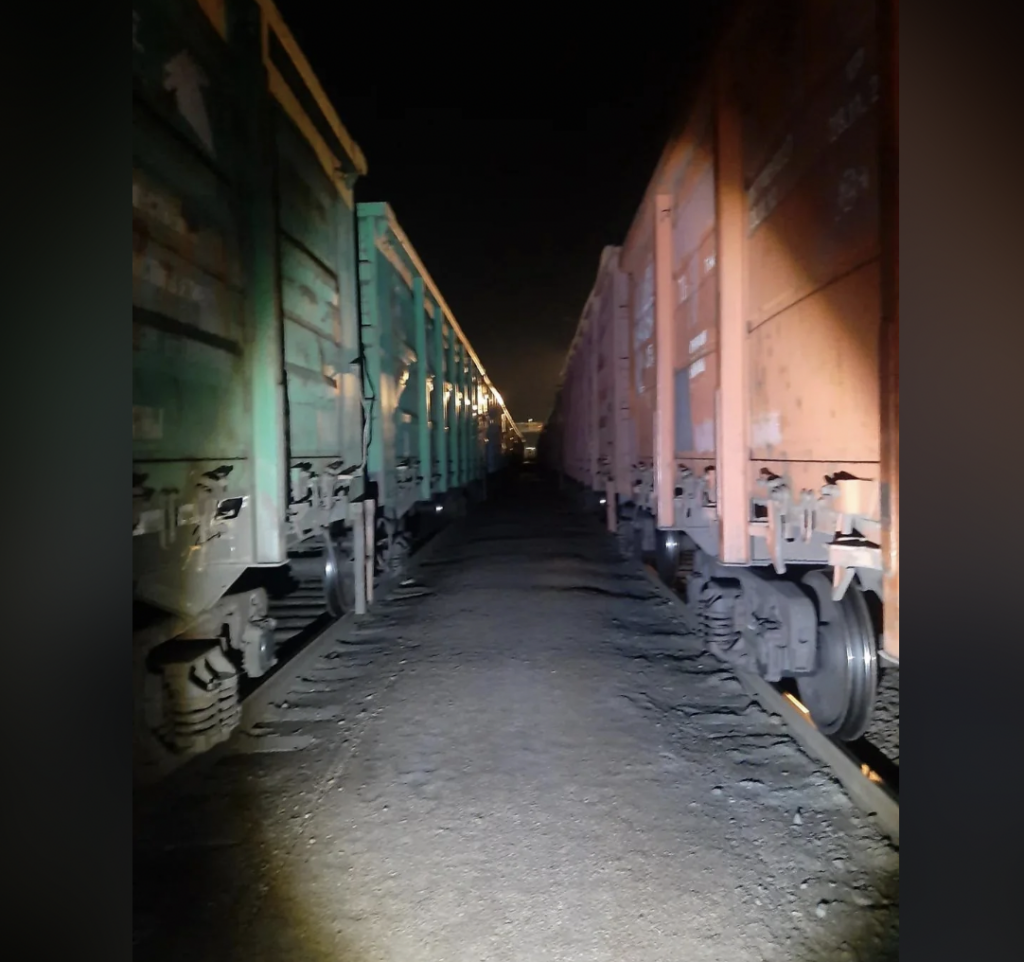 A night scene between two freight trains with green cars on the left and rusty orange cars on the right. The ground is covered in gravel, and the area is dimly lit, casting shadows on the train cars and tracks.