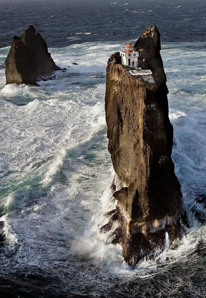 A dramatic coastal scene featuring a house precariously perched atop a tall, jagged sea stack, surrounded by turbulent ocean waves. The sky is overcast, and another rocky formation can be seen in the background.