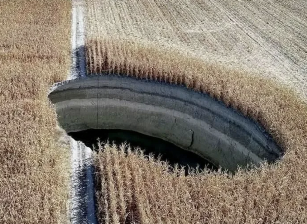 A large sinkhole in the middle of a cornfield, with the surrounding crops still intact. The image shows the gaping hole from an aerial perspective, highlighting the drastic disruption in the otherwise uniform, harvested field.