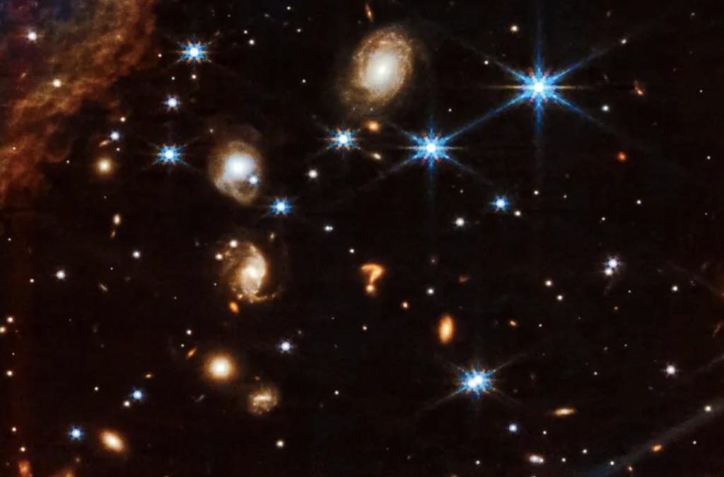A Hubble Space Telescope image shows a deep space view featuring several spiral galaxies, bright blue stars with cross-shaped diffraction spikes, and other glowing celestial objects set against a backdrop of countless distant stars.