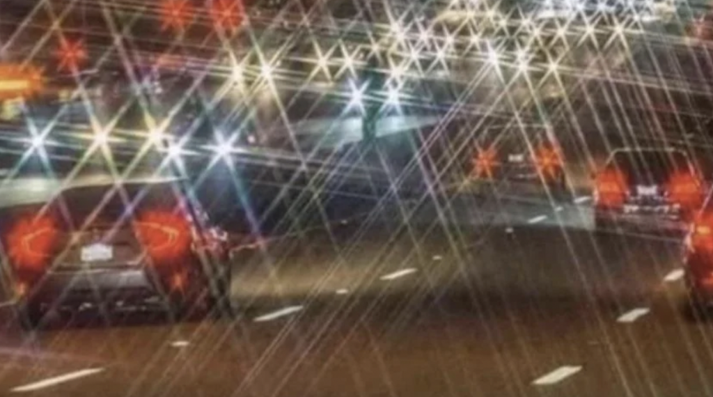 A nighttime highway scene shows cars with headlights producing star-shaped flares, likely due to rain or a special camera effect. The road is wet, reflecting light and creating a dazzling, crisscross pattern of bright streaks in the image.