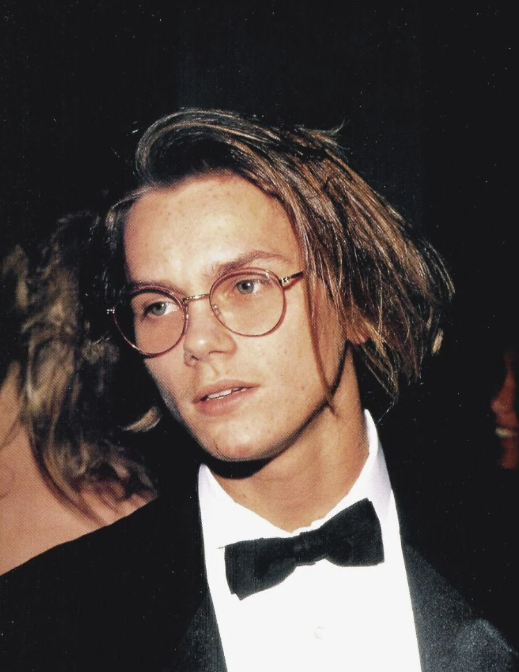 A young man with medium-length hair, wearing round glasses, a black tuxedo, and a bow tie. The background is dark, with a blurred figure visible behind him. He gazes slightly off to the side with a calm expression.