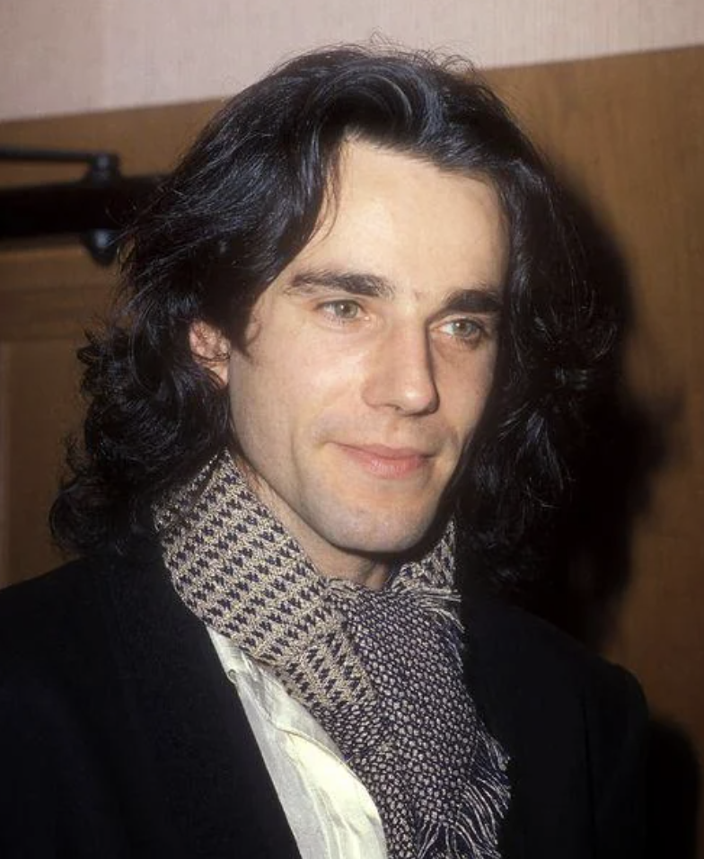 A man with shoulder-length dark hair and light skin is wearing a checkered scarf, a white shirt, and a dark jacket. He is looking slightly to the side with a neutral expression. The background is out of focus, showing a beige wall and possibly a wooden door.